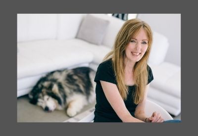 Image shows Rachel Amphlett posed in a studio laughing at the camera while the photographer's dog is sleeping on the floor behind her with its tongue out