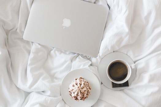 Image shows an Apple MacBook on a white duvet or counterpane next to a cup of coffee and a small cake on a plate
