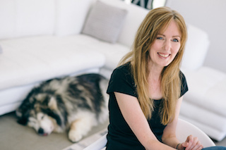 Image shows Rachel Amphlett posed in a studio laughing at the camera while the photographer's dog is sleeping on the floor behind her with its tongue out