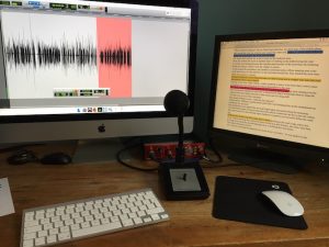 Image shows a studio microphone in front of a computer screen displaying audio waves with a book manuscript shown on the screen beside it