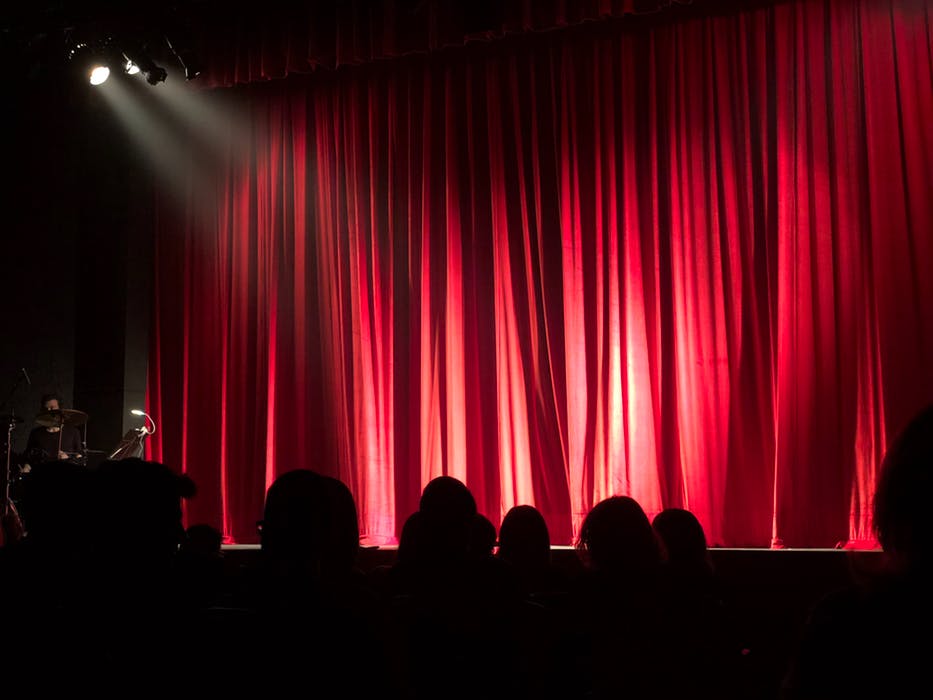 Image shows closed red theatre stage curtains with the silhouettes of an audience in front of the stage