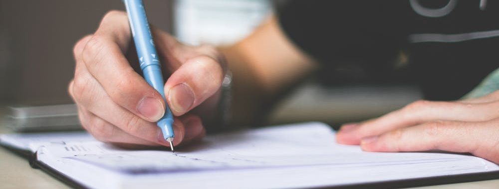 Image shows a woman writing in a notebook with a blue pen