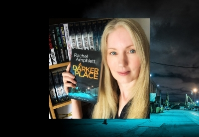 Rachel Amphlett holding a copy of A Darker Place next to a bookshelf displaying her books